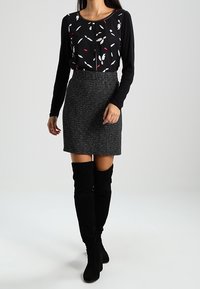 Schwarzes Langarmoberteil mit einem mehrfarbigen Blättermuster, kombiniert mit einem schwarzen Tweed-Minikleid und kniehohen schwarzen Wildlederstiefeln.