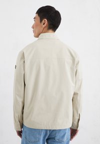 Light beige jacket with a collar and long sleeves, featuring a simple design and a patch on the left shoulder. Worn with blue jeans.