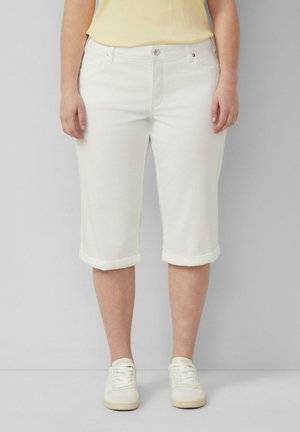 Frau trägt weiße, umgekrempelte Shorts bis zum Knie, hellgelbes ärmelloses Top und weiße Turnschuhe, steht vor einem einfarbigen grauen Hintergrund.