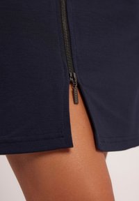 Granatowa spódnica z bocznym zamkiem błyskawicznym, wyposażona w metalową tasiemkę. Materiał jest gładki z delikatnym połyskiem i ma rozcięcie na dole dla większej swobody ruchów.