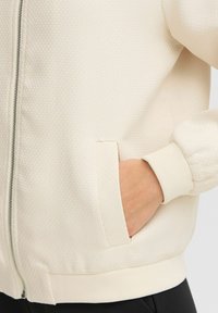 Cream-colored jacket with a textured fabric, featuring a full zipper, ribbed cuffs, and a side pocket. Simple and clean design.
