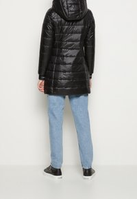 Persona di spalle che indossa un piumino con cappuccio nero, jeans azzurri chiari e sneakers nere con suole bianche.