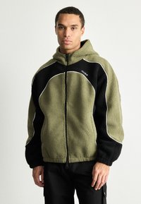 Fleece bundový svetr v olivově zelené a černé barvě, s otevřeným zipem, kapucí a bílými detaily. Měkká textilie s volným střihem.