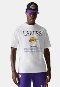 T-shirt grafico bianco con testo viola "LAKERS" e logo di un pallone da basket; abbinato a pantaloni felpati viola e un cappellino nero con accenti viola.