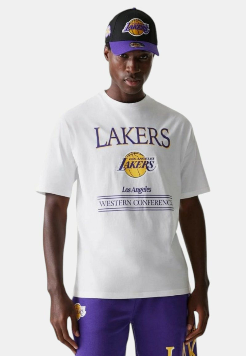 T-shirt grafico bianco con testo viola "LAKERS" e logo di un pallone da basket; abbinato a pantaloni felpati viola e un cappellino nero con accenti viola.
