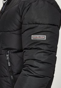 Schwarze Steppjacke mit wattierter Textur, ausgestattet mit einer Reißverschlusstasche und einem "KINGS WILL DREAM" Logo-Patch am Ärmel.