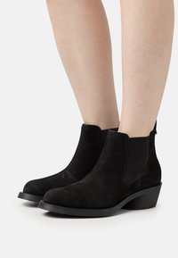 Botines de ante negro con punta redonda, paneles laterales elásticos y tacón bajo de bloque. Presentan una textura suave y costuras mínimas.