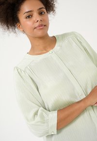 Blusa verde chiaro, texture con scollo rotondo, sottili righe verticali e maniche a sbuffo; presenta una chiusura con un solo bottone.