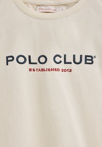 Kremowa bawełniana koszulka z okrągłym dekoltem, z haftowanym napisem "POLO CLUB" w kolorze granatowym oraz "ESTABLISHED 2012" w kolorze czerwonym z przodu.