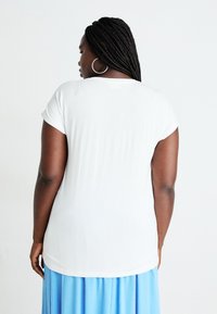 White short-sleeve t-shirt with a curved hem, smooth texture. Worn with a light blue pleated skirt, displaying a casual look from the back.