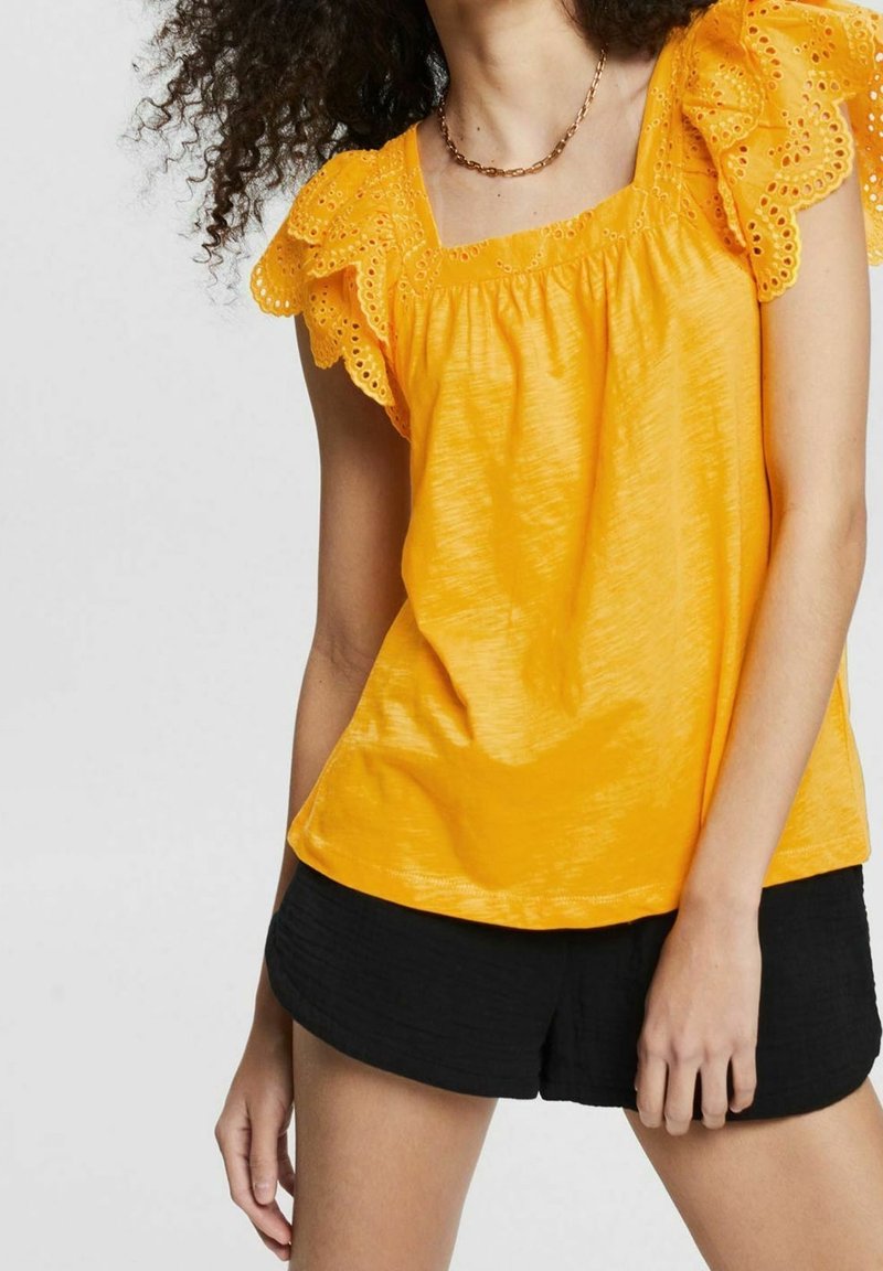 Femme portant un chemisier jaune vif avec des manches en dentelle ajourée et un short noir, debout avec un bras détendu et les cheveux bouclés visibles.