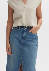Beige ärmelloses Oberteil mit V-Ausschnitt und Knöpfen, kombiniert mit einem blauen Jeansrock mit einem Schlitz vorne und einem schwarzen Gürtel.
