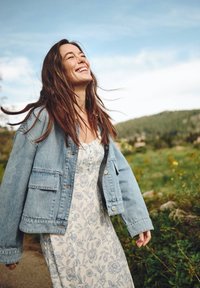 Jeansowa kurtka o luźnym kroju, jasnoniebieski kolor i duże kieszenie; noszona na białej sukience w niebieski kwiatowy wzór. Zielone otoczenie na zewnątrz.