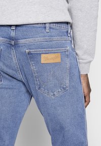 Detailní záběr na zadní kapsu modrých džínsů Wrangler, které jsou nošeny s šedým dlouhým rukávem, ukazující koženou nášivku značky a detaily šití.