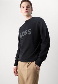 Zwarte sweatshirt met grijs "BOSS" logo, gekenmerkt door een ronde hals, lange mouwen en een zachte textuur, gecombineerd met beige op maat gemaakte broek.