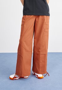 Baggy, rust-colored cargo pants with multiple pockets, drawstring hems, and large, rounded seams. Worn with white shoes featuring orange accents.