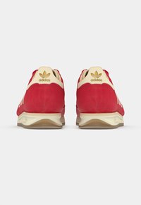 Červené tenisky s béžovými prvkami, semišový materiál, zaoblený tvar, biele pruhy a zlaté logo na päte. Plochá gumová podrážka.