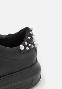 Baskets en cuir noir avec semelle en caoutchouc surélevée, ornées de pointes argentées sur le talon et d'un logo embossé. Texture lisse et forme arrondie.