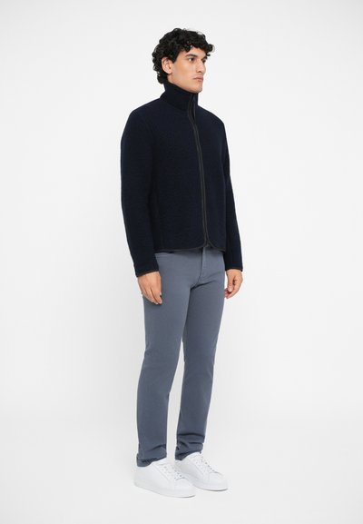 Marineblaue Fleecejacke mit hohem Kragen und Frontreißverschluss, kombiniert mit grauen Slim-Fit-Hosen und weißen Sneakers, an einem männlichen Model.