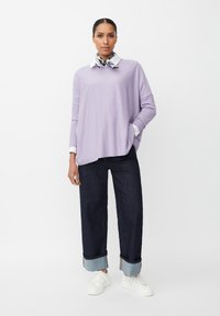 Maglione oversized lilla con maniche lunghe, camicia bianca con colletto sotto, abbinato a jeans scuri e sneakers bianche. Texture liscia.