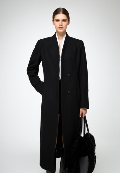 Cappotto lungo in lana nera con silhouette dritta, colletto a risvolto e chiusura a doppio bottone. Texture liscia, abbinato a una borsa nera.