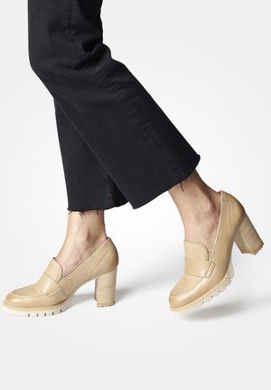 Voeten in beige leren loafers met blokhak, gecombineerd met zwarte cropped broek tegen een effen achtergrond.