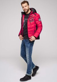 Chaqueta acolchada roja con detalles en negro, cierre de cremallera y capucha. Lleva puesta sobre un suéter oscuro y jeans azules con zapatos negros. Modelo posando.