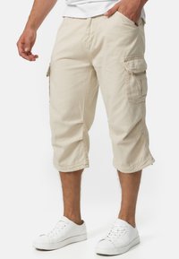 Shorts cargo beiges en coton, dotés de poches latérales et d'une coupe décontractée, avec des ourlets retroussés et une texture lisse. Portés avec des baskets blanches.