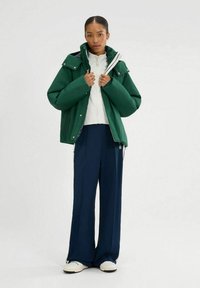 Zielona kurtka puchowa z wysokim kołnierzem, noszona na białej bluzie z zamkiem; zestawiona z granatowymi spodniami o szerokich nogawkach i białymi trampkami.