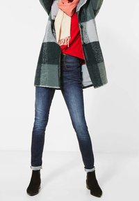 Rutig grön kappa över en röd topp, i kombination med mörka skinny jeans. Scarf är ljusrosa. Svarta anklarboots med låg klack.