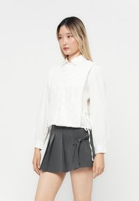 Hvit bluse med blonderdetaljer og knapper, sammen med et grått plissert skjørt med bånd. Stoffet er glatt, med en tettsittende silhuett.