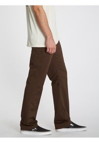 Homme portant un pantalon marron à jambes droites, des baskets noires slip-on et une chemise à manches courtes de couleur crème, debout de profil sur un fond blanc.