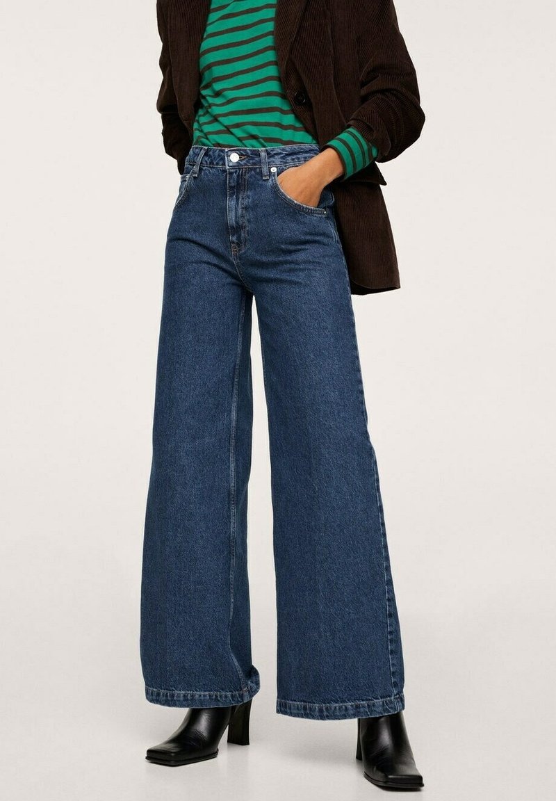 Žena v širokých tmavě modrých džínách, černých botách na podpatku, tričku s zeleno-černým pruhovaným vzorem a tmavě hnědém kabátu, stojí s jednou rukou v kapse.