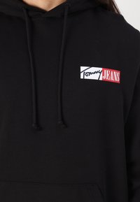Sort hætte sweatshirt lavet af blødt stof, med et fremtrædende frontlogo hvor "Tommy" er i hvid og "JEANS" i rød, snore ved halsen.