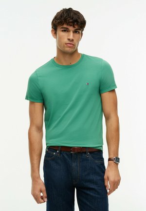 Ung mand iført en grøn kortærmet t-shirt, mørkeblå jeans, brun bælte og armbåndsur, stående mod en ensfarvet hvid baggrund.