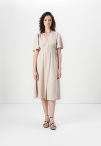 Abito midi beige chiaro con maniche a sbuffo corte, bottoni davanti e vita aderente. Indossato con sandali neri con cinturino su uno sfondo neutro.