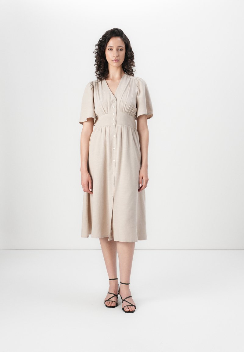 Robe midi beige clair avec des manches courtes à volants, une fermeture à boutons sur le devant et une taille cintrée. Portée avec des sandales noires à lanières sur un fond neutre.