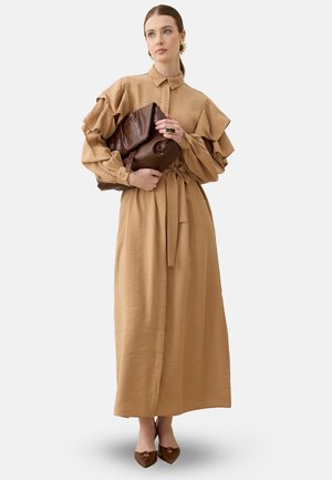 Frau trägt ein beige langärmliges, gerafftes Kleid und hält eine große braune, strukturierte Clutch, steht mit braunen spitzen Schuhen vor weißem Hintergrund.