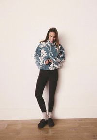 Sininen ja valkoinen kuvioitu fleece-pullove, jossa on korkea kaulus, yhdistetty mustiin legginsseihin ja ruskeisiin liukkaasti jalkaan puettaviin kenkiin. Puulattia taustalla.