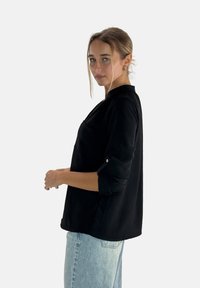 Maglietta nera a maniche lunghe con vestibilità rilassata, collo rotondo e tasca frontale. Polsini arrotolati e tessuto liscio; abbinata a jeans azzurri chiari.