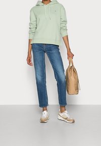 Sweat à capuche vert clair, jean droit bleu, baskets beige avec accents, et un sac à cordon beige tenu dans une main.