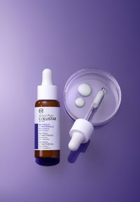 Bouteille en verre brun avec compte-gouttes blanc, étiquetée "Retinol + Panthénol." Accompagnée d'une assiette transparente avec deux petites gouttes brillantes.