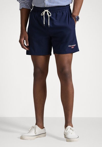 Marine svømmebukser med elastisk midje og snøring. Har "POLO SPORT" logo i rødt. Brukt med hvite sneakers.