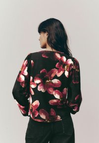 Blusa nera a maniche lunghe con grandi motivi floreali rosa, superficie testurizzata e maniche a sbuffo arricciate. Indossata con jeans scuri.