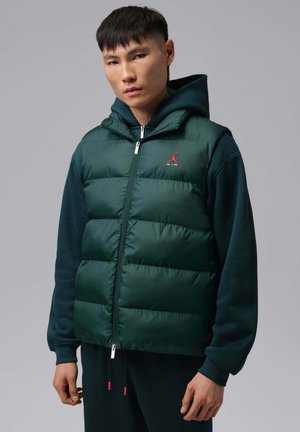 Un hombre luce un chaleco acolchado verde oscuro con cremallera sobre una sudadera con capucha, delante de un fondo gris liso de estudio.