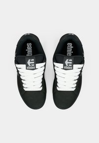 Schwarze Etnies-Sneaker mit weißen Schnürsenkeln, von oben betrachtet, wobei das Markenlogo auf der Zunge und der Innensohle sichtbar ist.