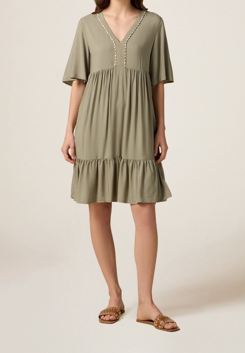 Robe vert olive à col en V avec manches courtes et larges ; taille froncée et ourlet à volants ; tissu doux avec un détail de couture subtil. Portée avec des sandales plates.