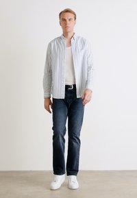 Gestreiftes Hemd in Hellblau und Weiß, kombiniert mit dunkelblauen Jeans und weißen Sneakers. Das Model steht vor einem weißen Hintergrund.