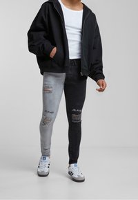 Vaqueros bicolor con pierna izquierda gris y pierna derecha negra, detalles desgastados en las rodillas, combinados con una sudadera negra y una camiseta blanca.