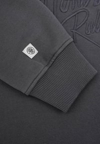 Grå sweatshirtdetalj med ribbad mudd, texturerad tyg och en fyrkantig vit etikett med logotyp. Broderad design, slät yta.